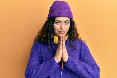 Young brunette woman with curly hair listening to music using headphones praying with hands together asking for forgiveness smiling confident. 