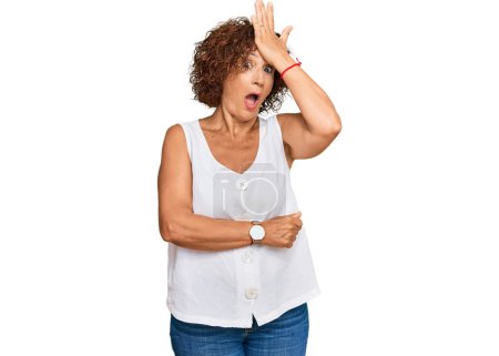 Foto de Hermosa mujer madura de mediana edad con camisa blanca casual sorprendida con la mano en la cabeza por error, recuerde el error. olvidado, mal concepto de memoria. - Imagen libre de derechos