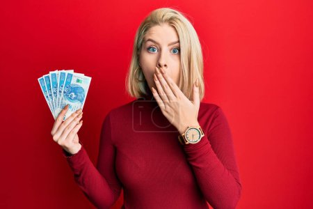 Joven mujer rubia sosteniendo 50 pulidos billetes zloty cubriendo la boca con la mano, conmocionada y temerosa de equivocarse. expresión sorprendida 