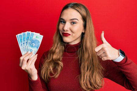 Joven mujer rubia sosteniendo 50 pulido zloty billetes sonriendo feliz y positivo, pulgar hacia arriba haciendo excelente y signo de aprobación 