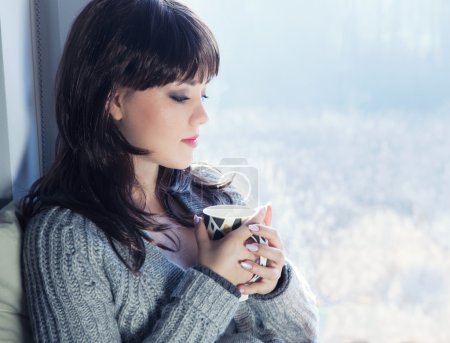 Téléchargez les photos : Jeune belle femme brune buvant du café et portant un cardigan tricoté assis à la maison relaxant par la fenêtre. Home chill out concept
. - en image libre de droit