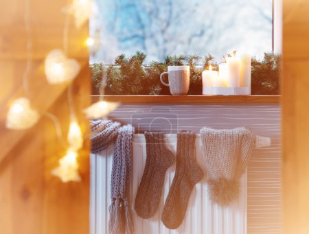 Téléchargez les photos : Accessoires de mode hiver chaussettes de chapeau séchant sur un appareil de chauffage, lumières de Noël, vue fenêtre de neige et boisson chaude - en image libre de droit