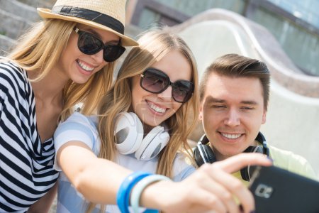 Foto de Tres jóvenes felices sentados al aire libre y haciendo selfies - Imagen libre de derechos