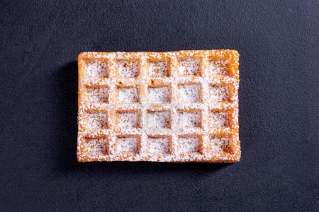 Sweet waffles sprinkled with powdered sugar prepared for serving. A tasty dessert eaten with fruit and preserves. Dark background.