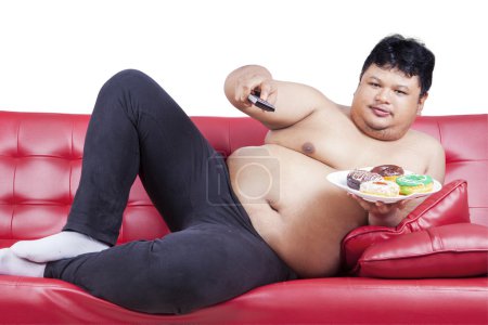 Photo for Photo of overweight man sitting on the sofa while holding a plate of donuts and remote, isolated on white background - Royalty Free Image