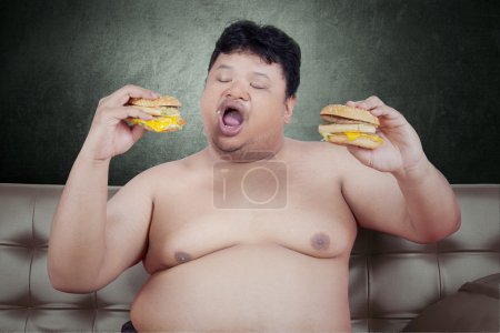 Foto de Foto de un joven obeso sentado en el sofá mientras come dos hamburguesas con queso - Imagen libre de derechos