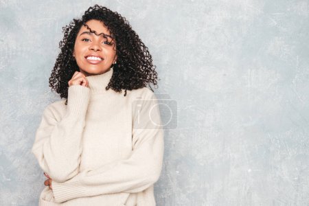 Retrato de una hermosa mujer negra con peinado afro rizos. Modelo sonriente en suéter blanco de moda. Sexy mujer despreocupada posando cerca de la pared gris en el interior del estudio. Bronceado y alegre 