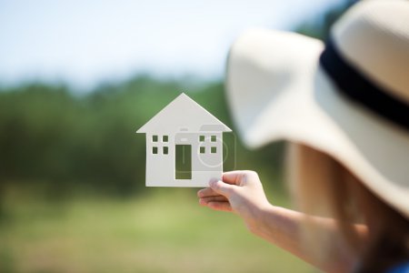 Foto de Mujer en sombrero de verano sosteniendo pequeña casa modelo al aire libre. Escena de naturaleza verde desenfocada en el fondo. Concepto inmobiliario, hipotecario, ecológico o de casa de campo - Imagen libre de derechos