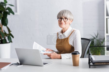 Téléchargez les photos : Mature chef d'équipe assis au bureau et tenant des papiers avec des graphiques et des graphiques dans le bureau - en image libre de droit