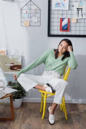 joven arquitecto afroamericano sentado en silla en el estudio en casa y mirando a la cámara