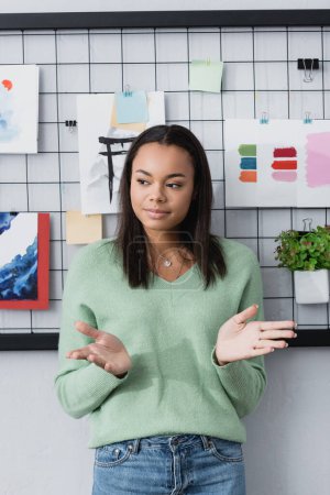 joven diseñador de interiores afroamericano gesto de pie cerca de pinturas en la pared