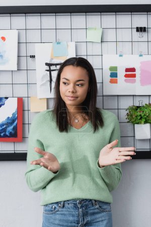 young african american interior designer gesturing while standing near paintings on wall