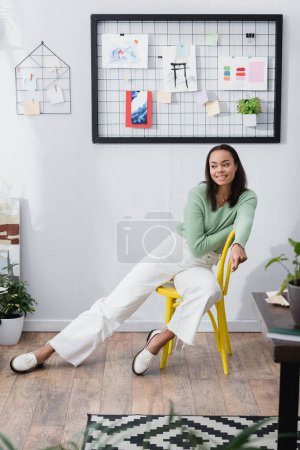 Foto de Sonriente diseñador de interiores afroamericano mirando hacia otro lado mientras está sentado en la silla en el estudio en casa, borrosa primer plano - Imagen libre de derechos