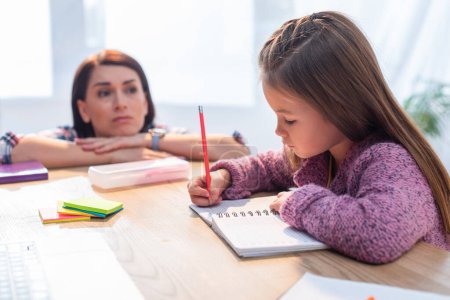Madre molesta mirando a su hija escribiendo en un cuaderno en el escritorio sobre un fondo borroso Madre molesta mirando a su hija escribiendo en un cuaderno en el escritorio sobre un fondo borroso