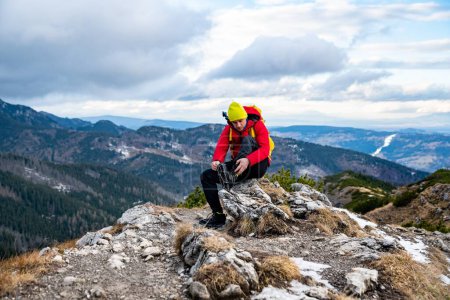 A tourist in a yellow cap and a red down jacket sits on a rock and puts crampons on his shoes.