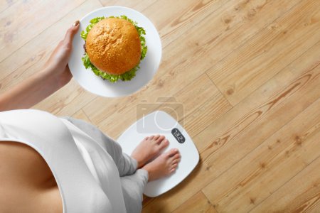 Dieta, comida rápida. Mujer con sobrepeso en escala, Hamburguesa. Comida chatarra
