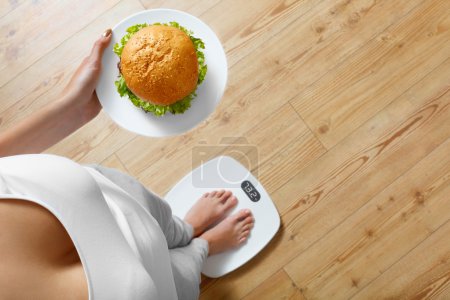 Téléchargez les photos : Diet And Fast Food Concept. Femme en surpoids debout sur l'échelle de pesée tenant Burger (hamburger). Malsaine malbouffe. Régime, mode de vie. Perte de poids. Obésité. Vue du dessus - en image libre de droit