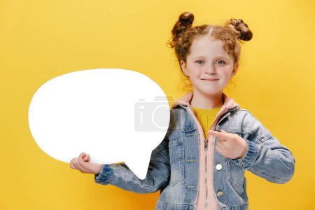 Portrait of cute little girl kid holding and pointing at blank speech bubble, wears denim jacket, isolated on yellow studio background wall with copy space for advertisement. People emotion concept