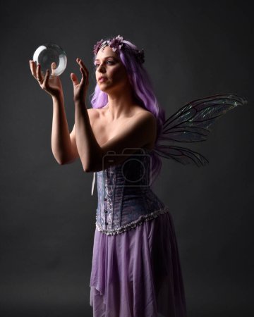 Close up portrait of a purple haired  girl wearing fantasy corset dress with fairy wings and flower crown, casting a spell.  Posing against a dark studio background with shadowed backlit lighting.