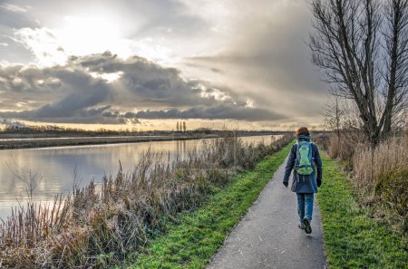 Téléchargez les photos : Maasland, Pays-Bas, 2 janvier 2021 : randonneur seul sur un étroit sentier asphalté le long d'un canal dans la réserve naturelle de vlietlanden sous un ciel spectaculaire un jour d'hiver - en image libre de droit