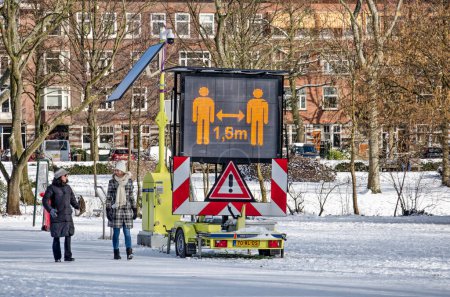 Téléchargez les photos : Rotterdam (Pays-Bas), le 10 février 2021 : deux personnes passent un panneau à matrice électronique dans le parc Vroesenpark, ce qui empiète sur la distance de 1,5 mètre comme mesure contre le covide 19 - en image libre de droit