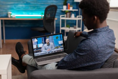 Téléchargez les photos : Étudiant afro-américain ayant webinaire de l'école de gestion en ligne sur ordinateur portable pendant le verrouillage du coronavirus dans le salon. Vidéocall réunion téléconférence télétravail sur ordinateur portable. Utilisateur informatique - en image libre de droit