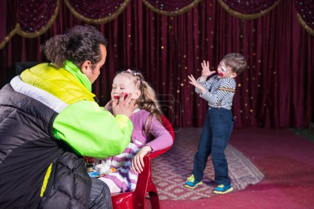 Téléchargez les photos : Homme appliquant le maquillage au visage de la fille blonde assise dans la chaise tandis que le jeune garçon clown effectue sur scène en arrière-plan - en image libre de droit