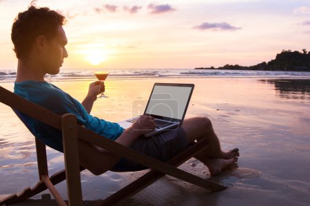 Foto de Joven hombre de negocios con cóctel trabajando en el ordenador portátil en la playa al atardecer, trabajo freelance en línea - Imagen libre de derechos