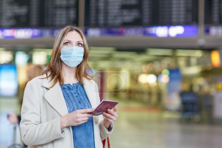 Mujer con máscara médica en el aeropuerto internacional, chequeando tablero electrónico y esperando vuelo. Pasajera con pasaporte europeo en terminal vacía debido a restricciones de viaje por coronavirus