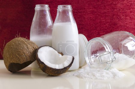 Foto de Productos de coco. Coco abierto agrietado con carne cortada por la mitad, hojuelas molidas en un frasco de albañil, harina y leche fresca en botellas de vidrio sobre una mesa con fondo de rubí rojo
. - Imagen libre de derechos