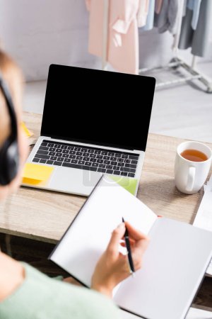 Laptop with blank screen near cup of tea and freelancer in headset writing on notebook on blurred foreground  