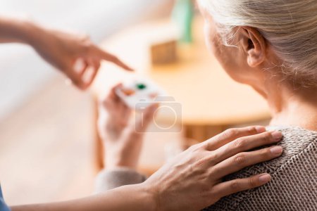 Teilaufnahme einer Krankenschwester, die die Schulter einer betagten Frau berührt und mit dem Finger auf Pillen auf verschwommenem Hintergrund zeigt