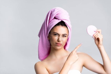 displeased beautiful woman with towel on hair pointing at sponge isolated on grey