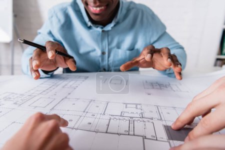 cropped view of african american businessman gesturing near blueprint and business partners on blurred foreground