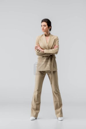 full length view of stylish woman looking away while posing with crossed arms on grey