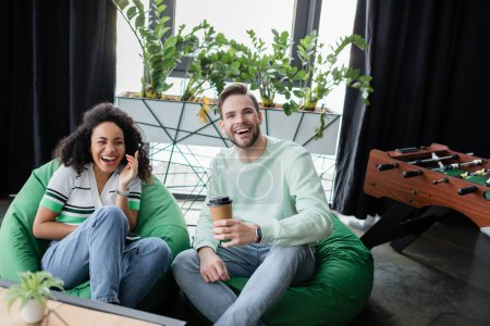 glücklicher Geschäftsmann mit Coffee to go, während er sich in der Büro-Lounge-Zone mit einem lachenden afrikanisch-amerikanischen Kollegen ausruht