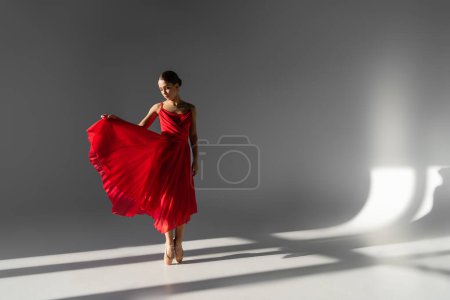 Ballerina in pointe shoes holding red dress on grey background with sunlight