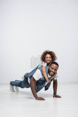 alegre africano americano chica piggybacking feliz papá haciendo flexiones en gris alegre africano americano chica piggybacking feliz papá haciendo flexiones en gris