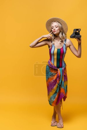 full length of blonde woman in straw hat pouting lips and holding vintage camera on yellow