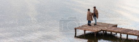 Young interracial couple with cups standing on pier near lake, banner 