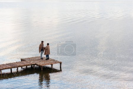 Pareja multiétnica en mantas sosteniendo copas mientras habla en el muelle 