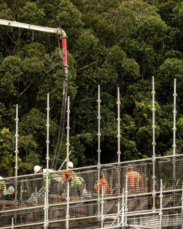 Foto de Los trabajadores cierran la diseminación de hormigón entregado por Boom Pump en un nuevo sitio de construcción con telón de fondo de arbustos. Nueva vivienda social en 56-58 Beane St. Gosford. En Australia. 17 de abril de 2021. Parte de una serie. Imagen comercial. - Imagen libre de derechos