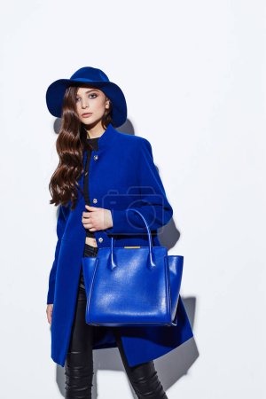 High-Fashion-Porträt einer jungen eleganten Frau. Blauer Mantel, Hut, Tasche. Weißer Hintergrund