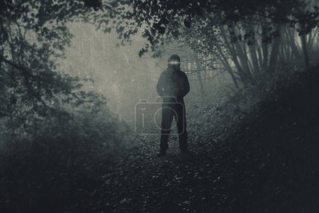 A scary hooded figure with glowing eyes on a path through a moody misty autumn woodland. With a grunge, blurred, textured edit