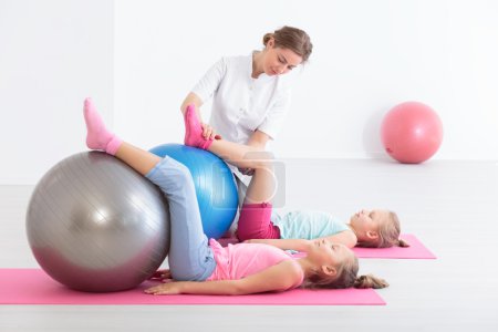 Photo for Two school girls lying on exercise mats exercising their legs on balls, accompanied by a physical therapist - Royalty Free Image