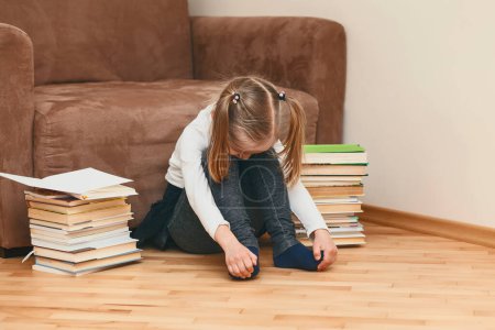 Téléchargez les photos : L'enfant s'ennuie assis sur le sol. La petite fille en a marre de l'école à domicile. Enfant fatigué à la maison sur un fond de livres - en image libre de droit