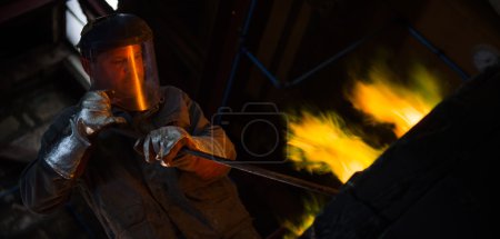 Photo for Steel worker in protective clothing raking furnace in an industrial foundry - Royalty Free Image