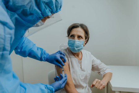 Foto de Médico varón en general protector, protector facial, máscara y guantes haciendo la vacunación COVID-19 para trabajadora de la salud mujer madura en el hospital. - Imagen libre de derechos