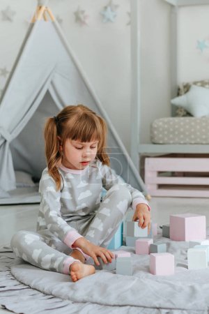 Hermosa niña pequeña con pijamas jugando con bloques de juguete de madera en la habitación de los niños. Concepto de juguetes ecológicos naturales. Enfoque selectivo.