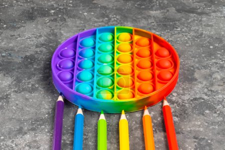 Foto de Juguete de silicona infantil antiestrés pop it o simple hoyuelo con lápices de todos los colores del arco iris sobre un fondo oscuro - Imagen libre de derechos